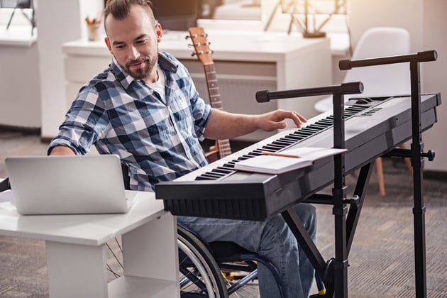 A white man in a wheelchair is indoors, performing in what appears to be his home on a keyboard, while leaning over to type something into his laptop.
