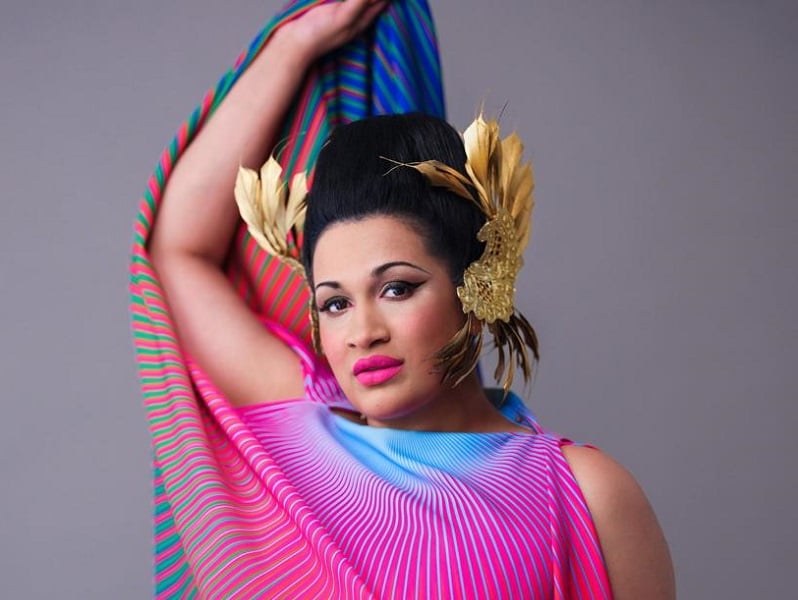 Photograph of Bishi Bhattachaya, she is wearing a brightly coloured top and gold head piece, and is looking directly into the camera with one arm raised.