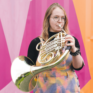 A photo of Lynn Henderson playing a French horn against a colourful background.
