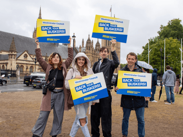 Four people standing with 'Back your buskers' plackards with the Westminster building in the background