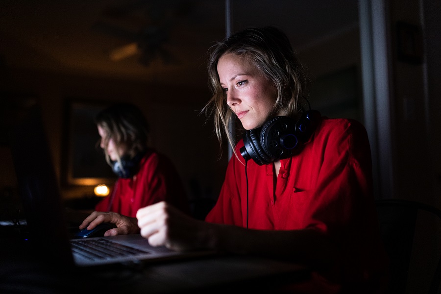 Woman with blonde hair in red shirt sat in very dark room. Her headphones are around her neck and she is illuminated by the open laptop screen.