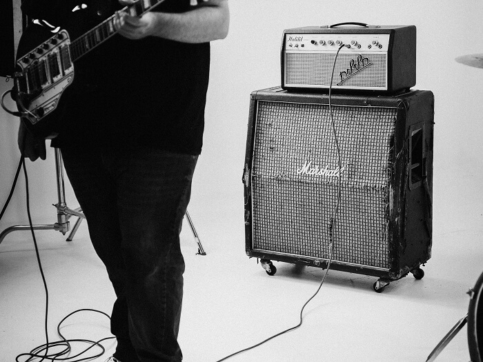 In black and white we see a persons legs, holding a guitar and standing in front of a large amplifier.