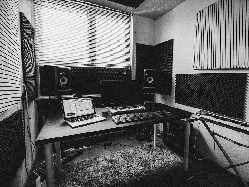 Black and white photograph of someone's home recording studio set up, two computers and a keyboard are on a desk and the walls are sound proofed.