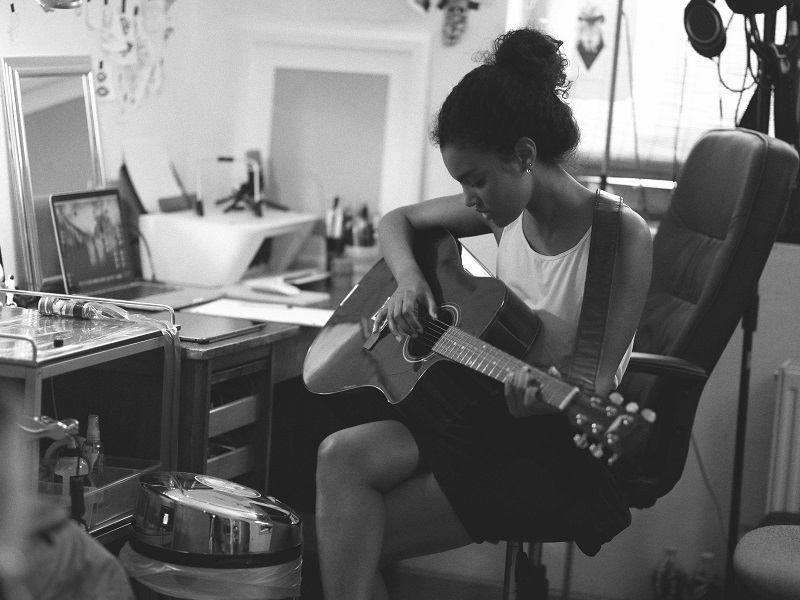 Black and white photograph of a woman playing an acoustic guitar in front of a laptop, in what appears to be a bedroom.