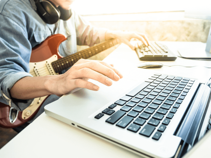Close up of musician wearing headphones with an electric guitar on their knee, they are typing on a keyboard in front of an open laptop.