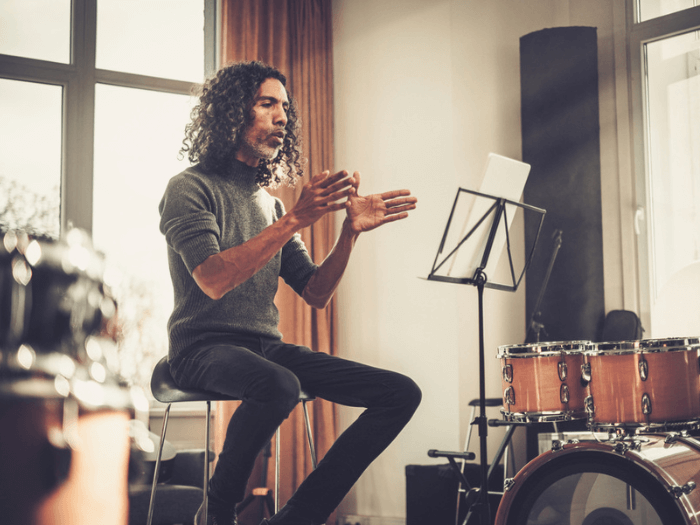 Male music teacher, on a stool in front of class and a drum kit, explaining something visually with their hands.