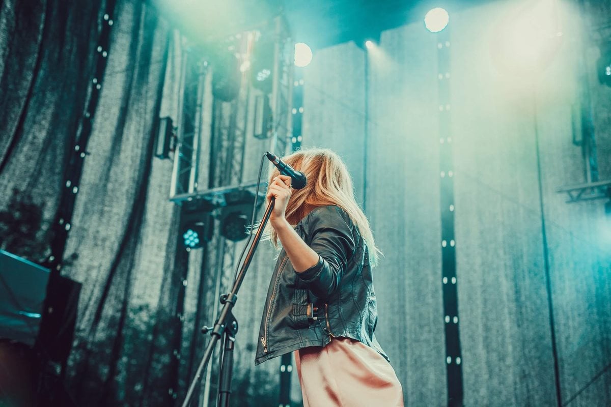 Photograph of a woman on stage, she's holding a microphone in one hand and looking towards the stage back. We can't see her face, which is hidden behind a curtain of hair.