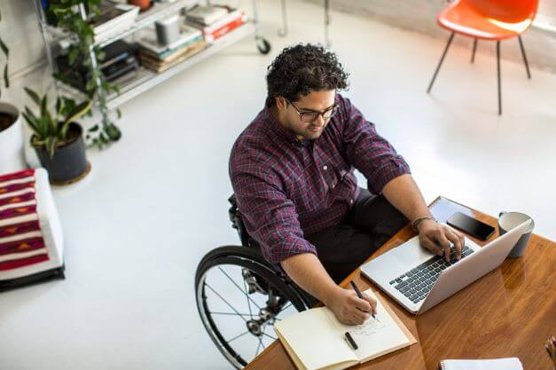 a man in a wheelchair at his desk, working on a laptop and making notes