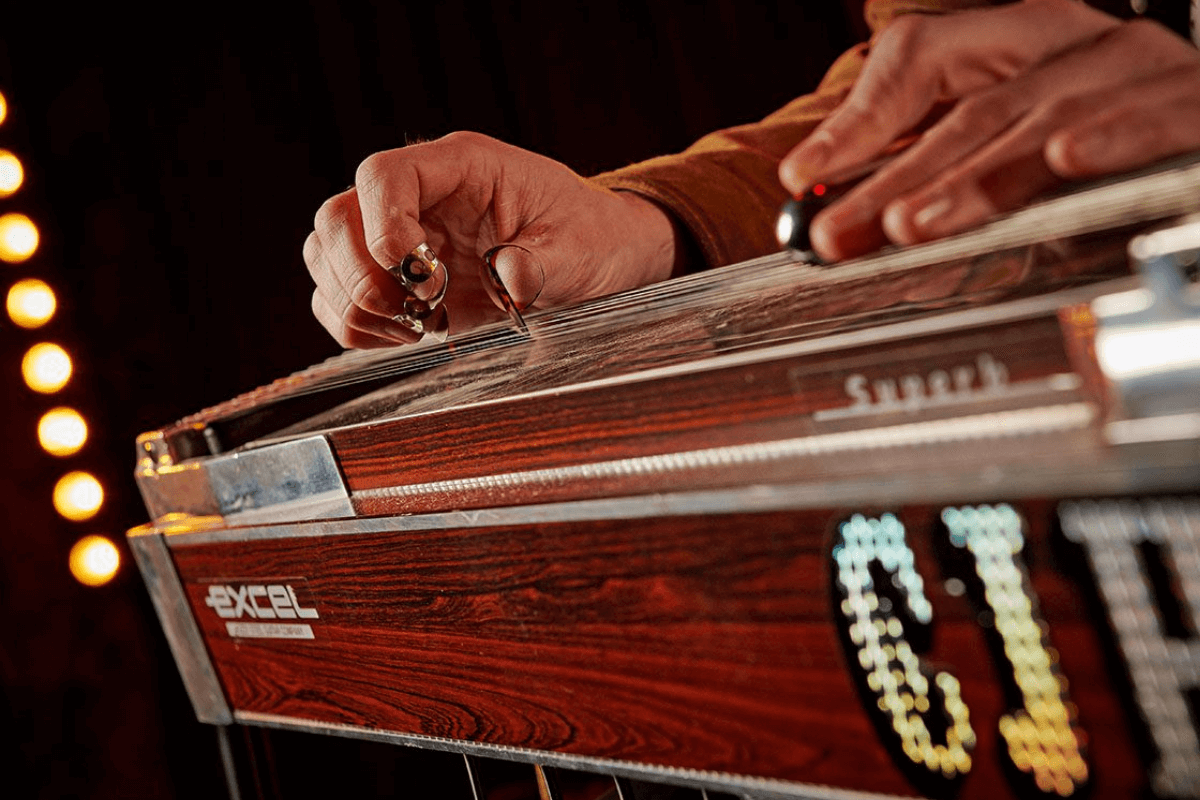 Close up of CJ's hands playing steel guitar with pics on fingers against a lit up dark background.