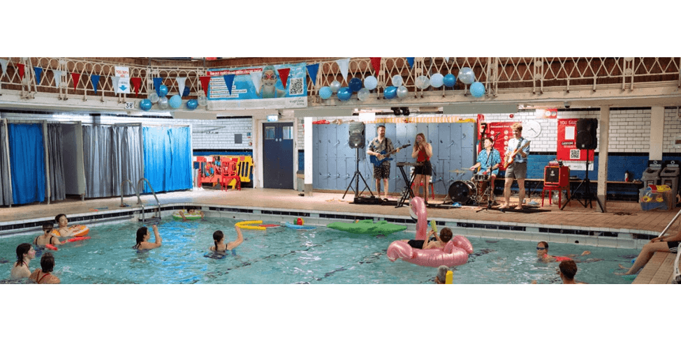 Poppy and her band at the poolside performing live. People are in the water, in rings and inflatables watching. Banners and balloons are behind the band.