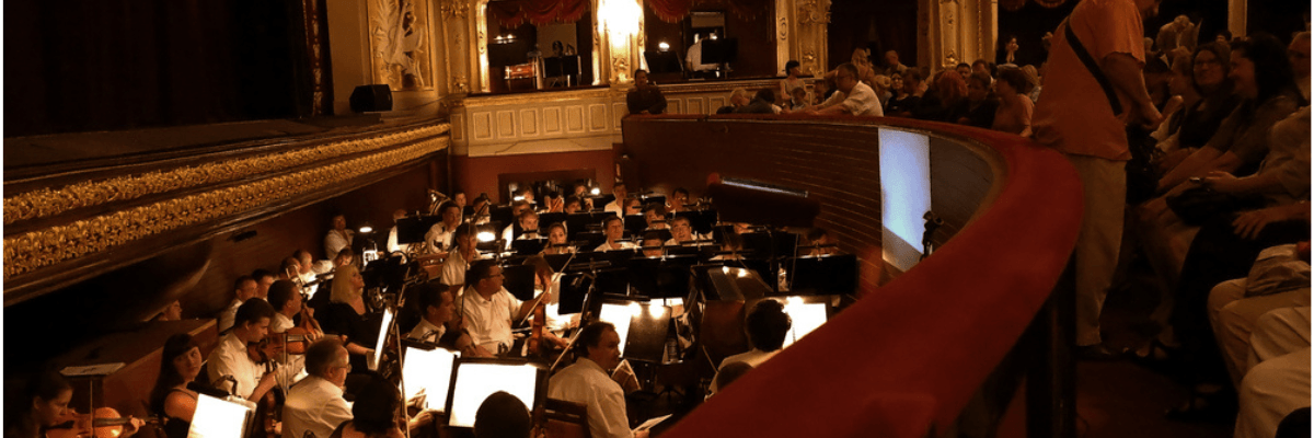 A theatre pit at the front bottom of the stage, full of musicians lit by lamps, as the audience takes their seats before the performance.