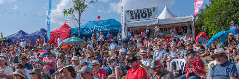 Large crowd sat in front of the main stage at Tolpuddle. It is a sunny day and there are lots of festival tents.