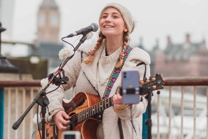 A woman is busking with a guitar and microphone on a busy London street, with a phone set up to take contactless payments