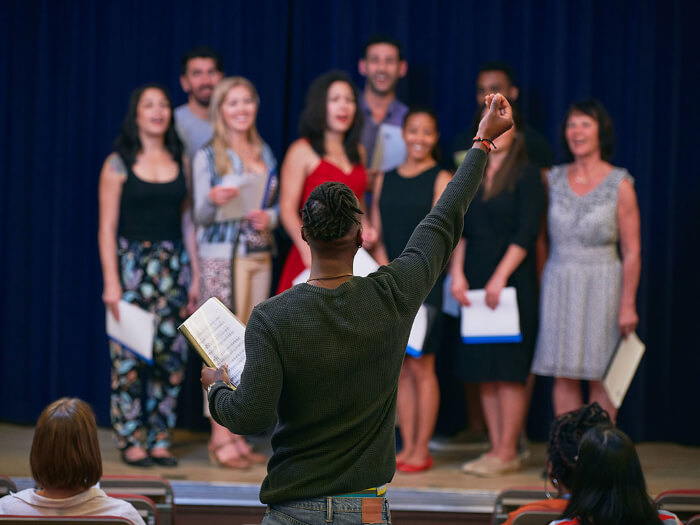 A conductor raises a hand towards a choir stood on a stage, facing outwards they appear to be singing.