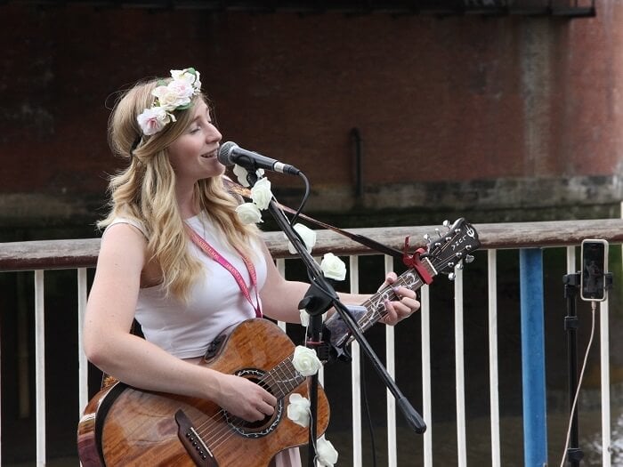 Busker Charlotte Campbell, a white woman with long blonde hair sings in an outdoor location, smiling into the microphone and strumming an acoustic guitar.