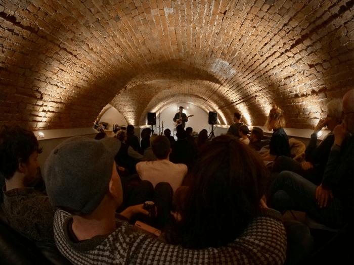 Man performing on stage with guitar under a brick arch/tunnel way, the crowd are sat watching.