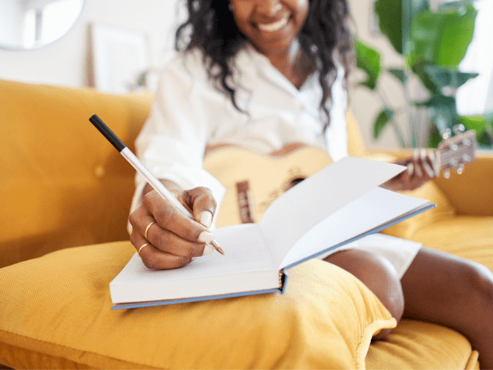 Black women sat on sofa, smiling as she balances an acoustic guitar on her knee and writes in a notebook with a pen