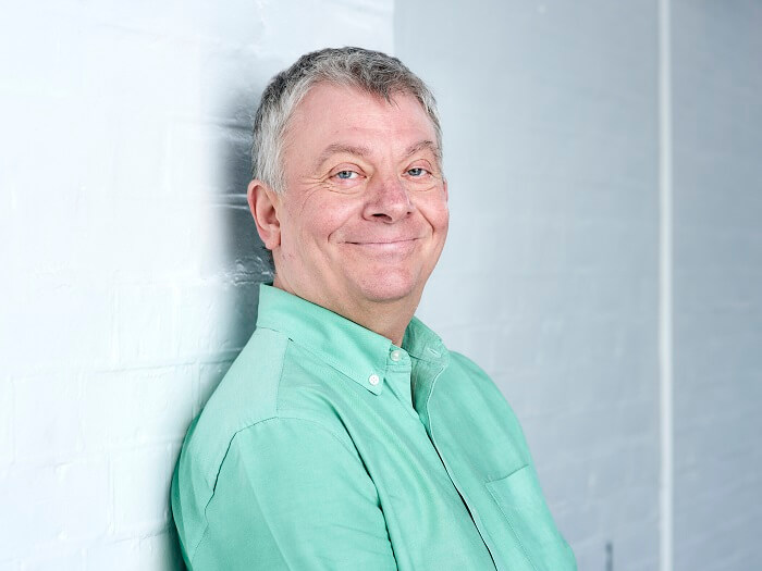 Alex Gascoine, a white man with grey hair and a brightly coloured mint green short is leaning against a white painted wall and smiling into the camera.