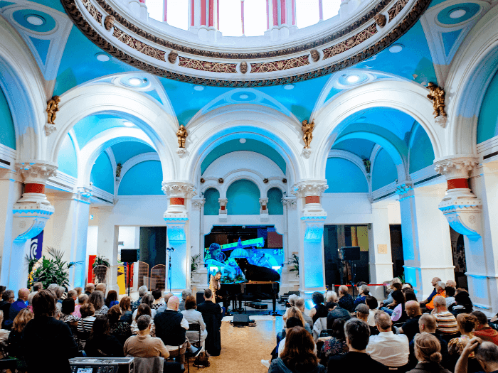 A photo of an audience sitting in a room with a high domed ceiling with intricate decoration.