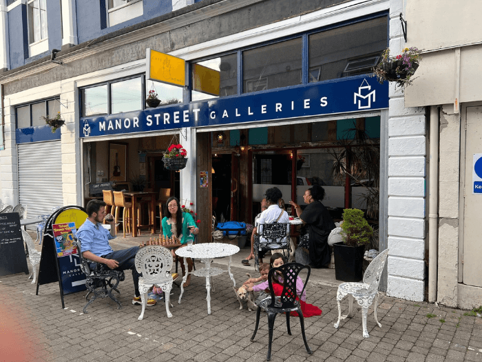 Day time street view of the venue with its doors open, people are sat talking and having a drink.