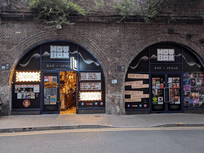 Exterior of the venue, nestled under two stone archways, with black windows. Posters and lit up signs cover the front.