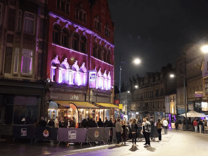 Exterior shot of the venue at night, lit up on a highstreet. Lots of people are standing around outside, queuing to get in, and in the smoking area.