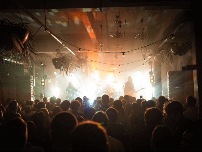 Inside of the smaller venue room, a band is playing on stage to the crowd, with lights, plants and chandelier's handing from the ceiling.