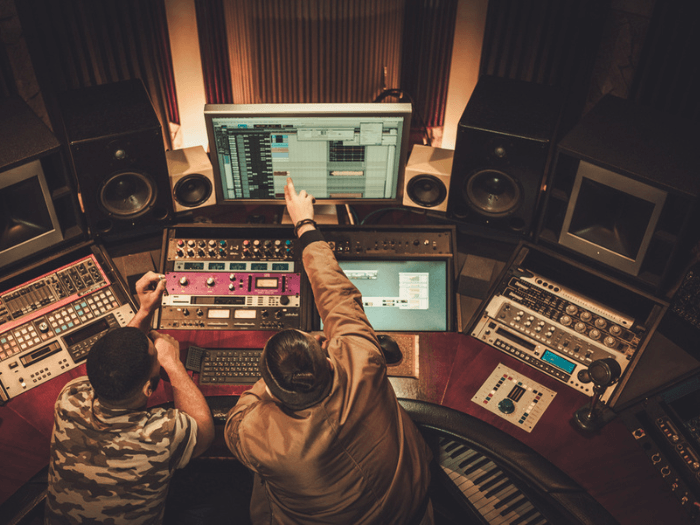 Birdseye view of two people sat in a recording studio, surrounded by controls and keyboards, pointing to a computer screen.
