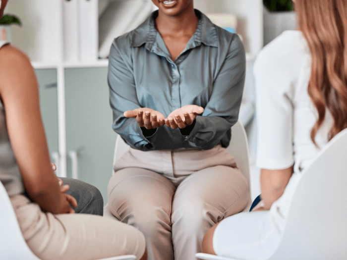 Three people sat in a circle, the person speaking has their arms open with thier palms facing upwards, representing transparency.