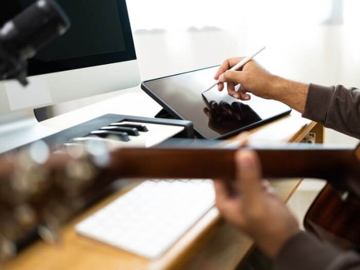 A person sat in front of a computer with guitar, keyboard, and tablet - which they are writing music on.