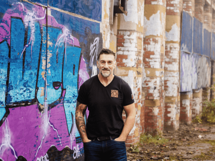 Dene Josham smiling and standing in front of a colourful blue and purple graffitied wall.