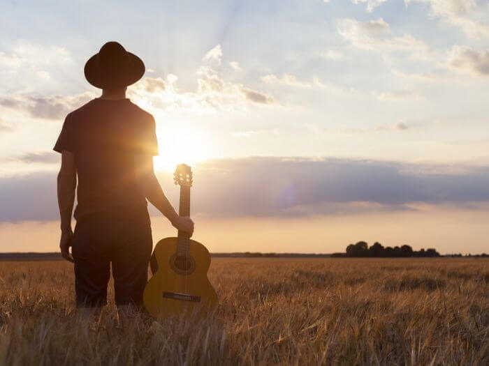 Shiloutte of a person standing in a field, wearing a hat, holding a guitar at sunset.
