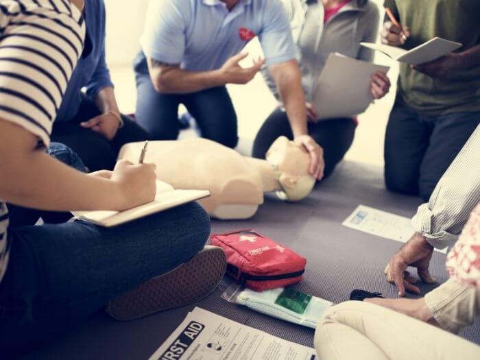 People sat around a first aid training manikin taking notes as the person demonstrates.