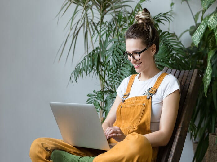 Woman sat cross legged in yellow dungarees, smiling as she looks at an laptop on her knees.