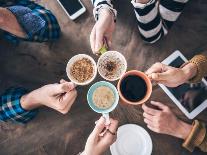 Birdseye view of four people 'cheers-ing' with cups of tea and coffee, laptops and phones are on the desk.