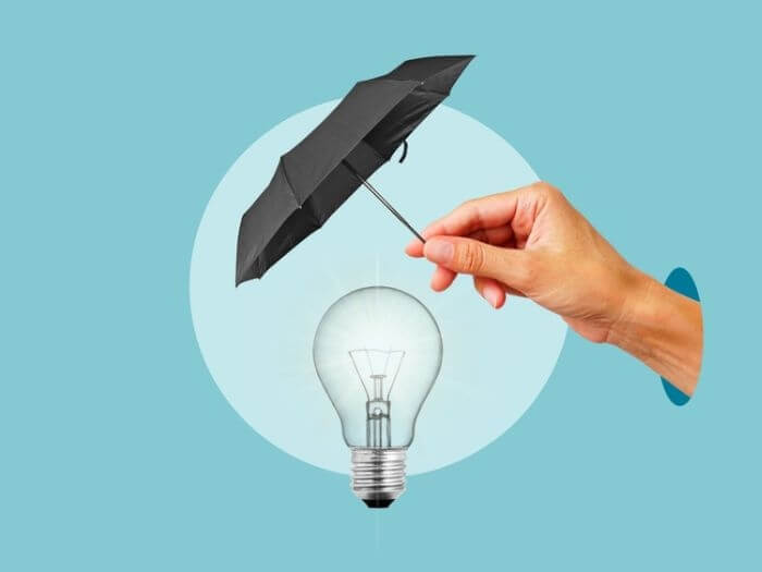 Hand holding a small black umbrella over a lightbulb on a blue background. Protection of Intellectual Property concept.