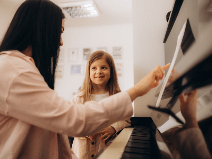 A woman sat at a piano, smiling as she teaches a young girl how to play.