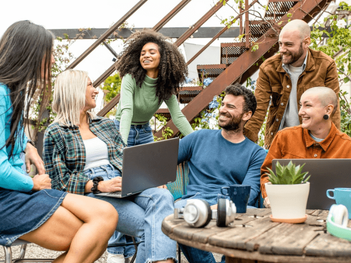 A diverse group of people, sat around an outdoor table smiling with laptops and mugs.