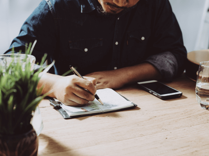 Person sat at a table with their phone and glass of water. sat with some paper making notes with a pen.