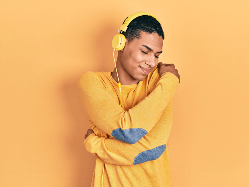 Man in yellow jumper and yellow background with headphones on smiling with his arms wrapped around himself.