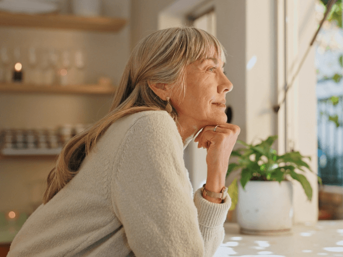 An older lady, chin propped on hand, at home smiling into her sunny garden, candles and a calming background behind her.