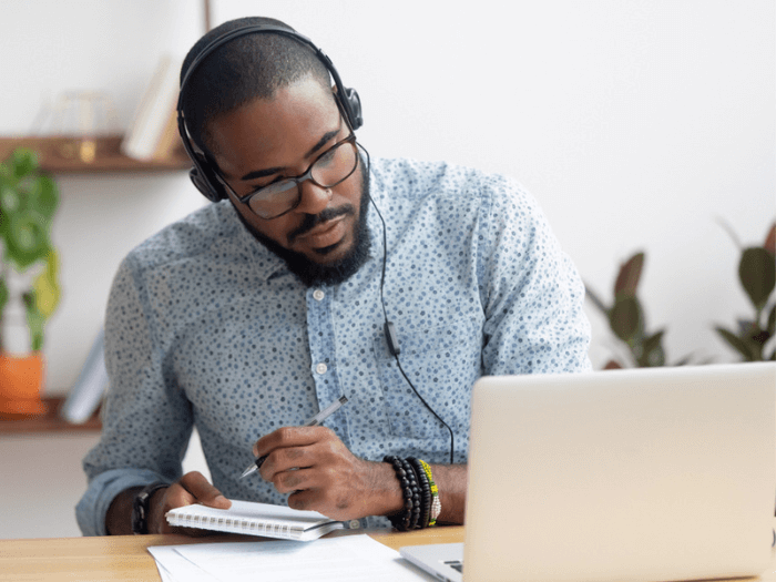 Young Black male wearing headphones and glasses, taking notes whilst watching a laptop screen.