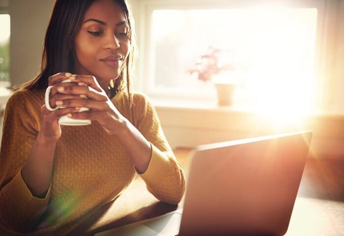 A woman sat in front of an open laptop smiling, holding a mug. Sunlight streams in through the window.
