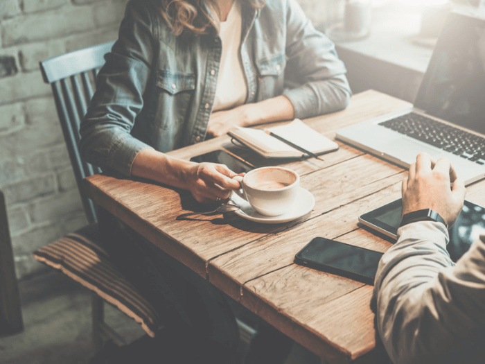 Two people sat having a coffee in a coffee shop, with laptop, phones and notebooks.