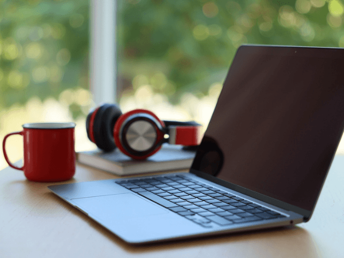 Open laptop, mug, headphones and book on a wooden table against a blurred green, outside background, closeup.