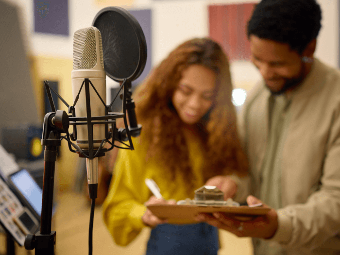 Two people in the background of a recording studio, writing something on a clipboard, a mic with a pop shield is in the foreground.