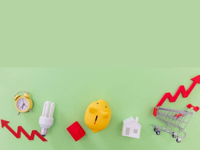 Cost of living concept. Flat lay of a piggy bank, light bulb, shopping trolley and red arrows going up and down against a green background.