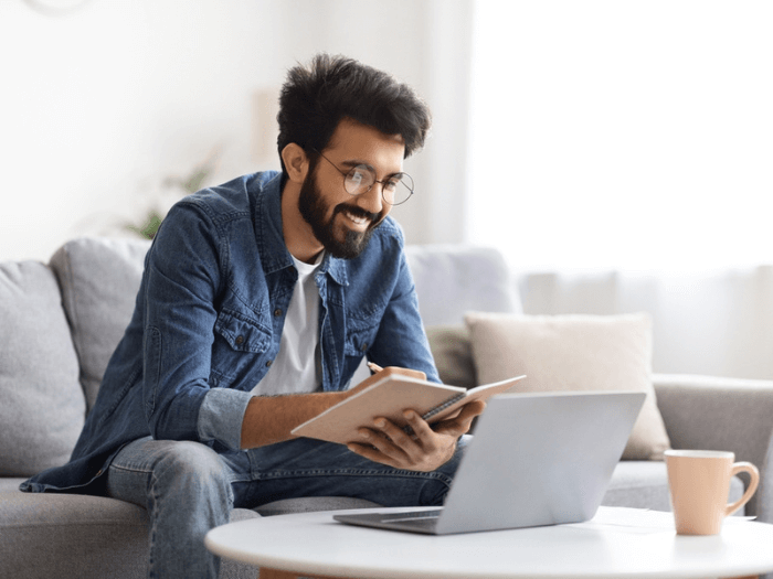 Man sat at a laptop at home smiling, sitting on the sofa with a mug, making notes in a book.