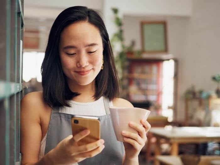Young woman smiling looking at the phone in her hand while holding a mug in the other.