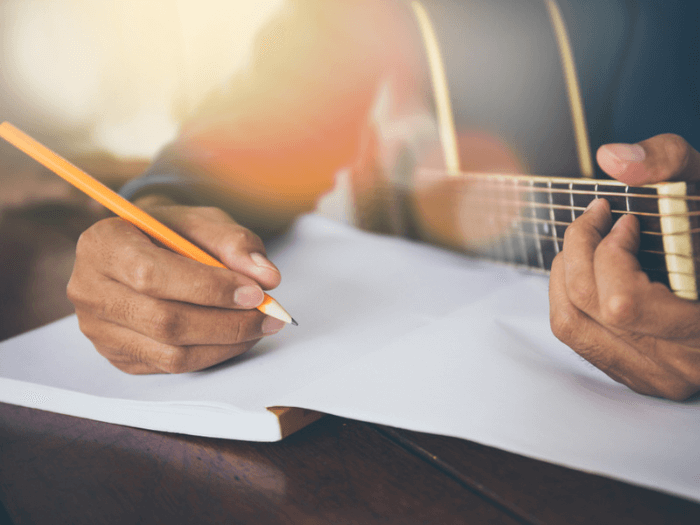 Close-up of a hand holding a pencil writing in a blank notebook, while the other hand holds a chord on an acoustic guitar.
